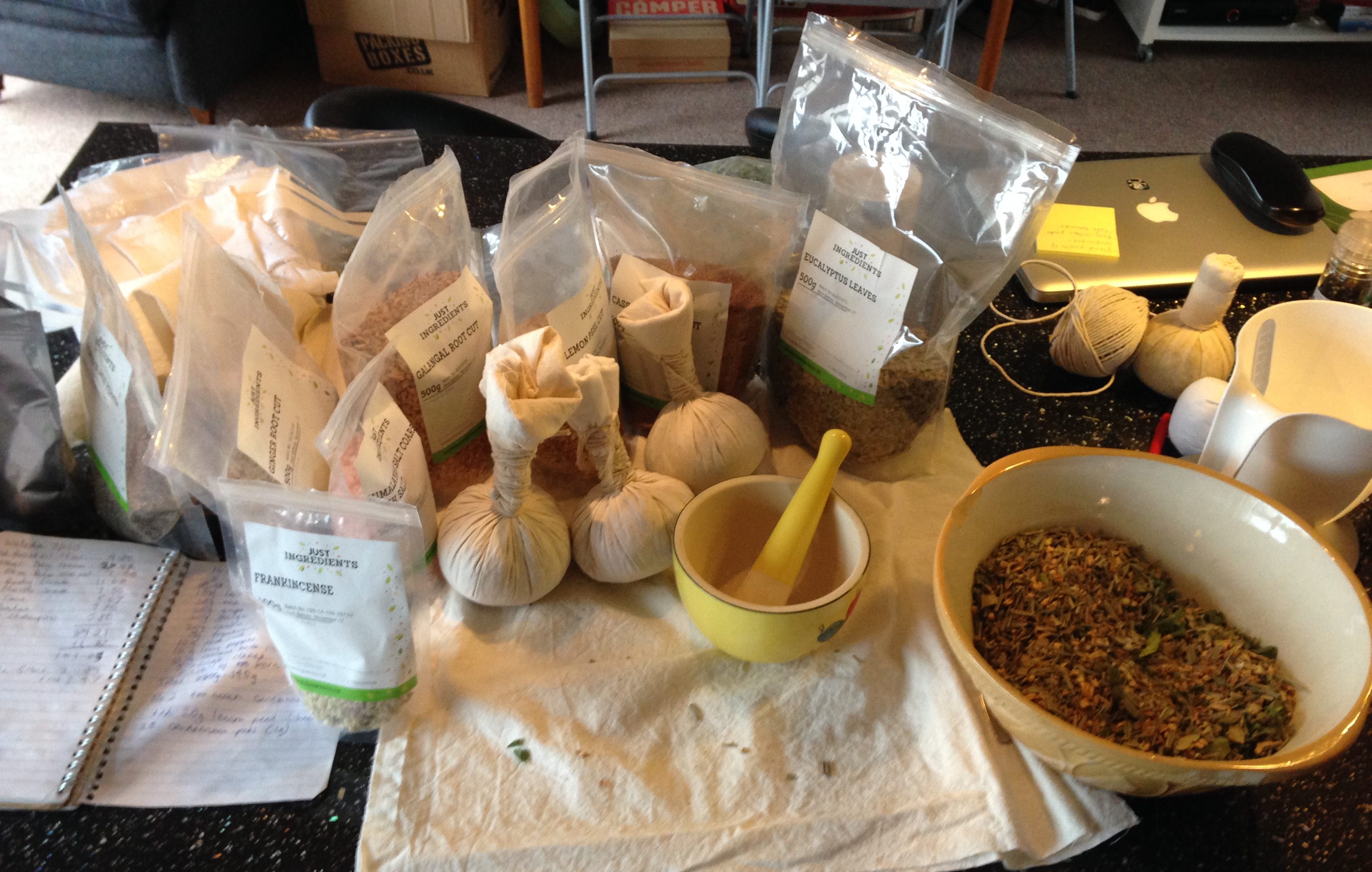 An arrangement of the ingredients to make a hot herbal compress, including a selection of herbs in containers, a bowl of herbs, a pestle and mortar, some hot compresses already made, a notebook, and a cloth